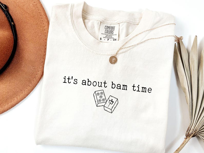 Pu&ograve; includere: T-shirt color crema con la scritta nera "it's about bam time". La maglietta presenta una grafica di due tessere di mahjong. Una collana d'oro con un ciondolo &egrave; drappeggiata sulla maglietta.