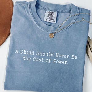 Puede incluir: Una camiseta azul Comfort Colors con el texto "A Child Should Never Be the Cost of Power". Un collar dorado con un colgante descansa sobre la camiseta. Un sombrero marr&oacute;n es visible en el fondo.
