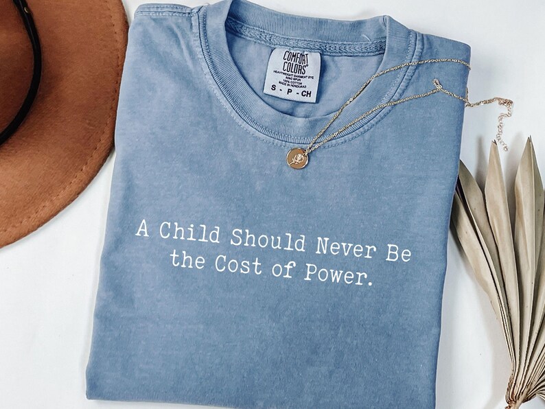 Puede incluir: Una camiseta azul Comfort Colors con el texto "A Child Should Never Be the Cost of Power". Un collar dorado con un colgante descansa sobre la camiseta. Un sombrero marr&oacute;n es visible en el fondo.