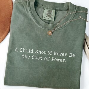 Puede incluir: Camiseta Comfort Colors verde oliva con el texto "A Child Should Never Be the Cost of Power". Un collar dorado con un colgante descansa sobre la camiseta. Un sombrero marr&oacute;n y una planta seca est&aacute;n en el fondo.