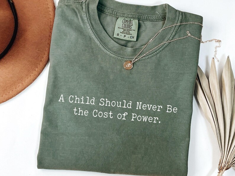 Puede incluir: Camiseta Comfort Colors verde oliva con el texto "A Child Should Never Be the Cost of Power". Un collar dorado con un colgante descansa sobre la camiseta. Un sombrero marr&oacute;n y una planta seca est&aacute;n en el fondo.