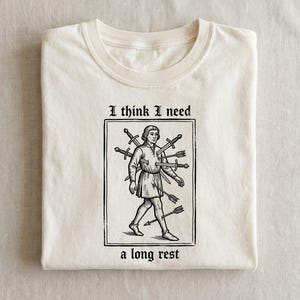 May include: Cream-colored t-shirt with a black and white illustration of a person with swords and arrows. The text reads "I think I need a long rest". The shirt is folded neatly.