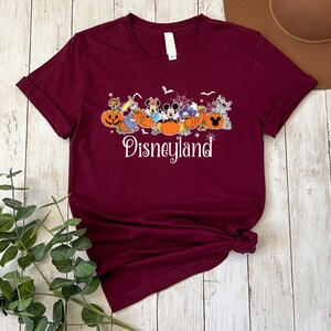 May include: A maroon t-shirt with a graphic of Mickey Mouse and friends dressed in Halloween costumes. The graphic includes pumpkins, bats, and a spider web. The text "Disneyland" is printed below the graphic.