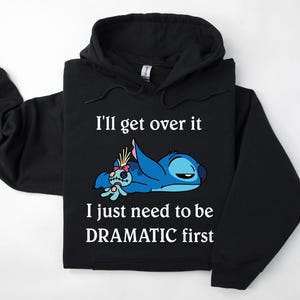 May include: Black hoodie featuring a blue Stitch character from Lilo & Stitch lying down with a small doll. The text on the hoodie reads "I'll get over it I just need to be DRAMATIC first."