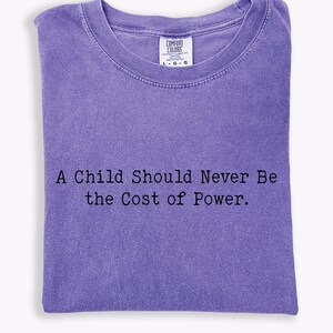 Puede incluir: Una camiseta Comfort Colors de color lavanda con el texto "A Child Should Never Be the Cost of Power". La camiseta est&aacute; doblada y tiene cuello redondo. La camiseta tiene una etiqueta en el cuello.