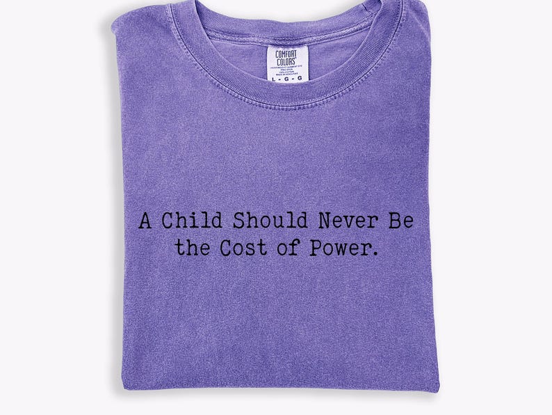 Puede incluir: Una camiseta Comfort Colors de color lavanda con el texto "A Child Should Never Be the Cost of Power". La camiseta est&aacute; doblada y tiene cuello redondo. La camiseta tiene una etiqueta en el cuello.