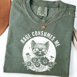 Puede incluir: Camiseta verde oliva con el texto "RAGE CONSUMES ME" que rodea una ilustraci&oacute;n de un gato. El gato est&aacute; en un c&iacute;rculo blanco con flores. Un collar dorado con un peque&ntilde;o colgante est&aacute; sobre la camiseta.