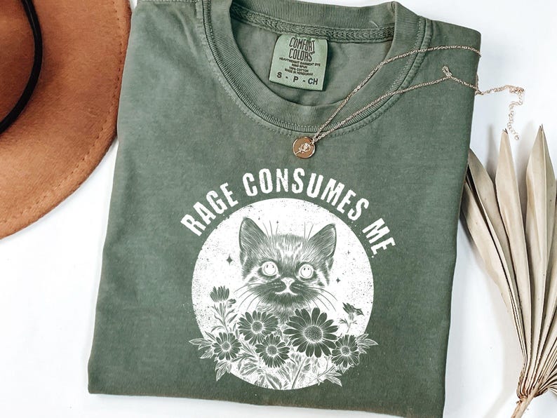 Puede incluir: Camiseta verde oliva con el texto "RAGE CONSUMES ME" que rodea una ilustraci&oacute;n de un gato. El gato est&aacute; en un c&iacute;rculo blanco con flores. Un collar dorado con un peque&ntilde;o colgante est&aacute; sobre la camiseta.