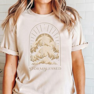May include: A beige t-shirt with a graphic of a stylized cloud with a sun and rays behind it. The text "STORMBLESSED" is printed below the graphic.