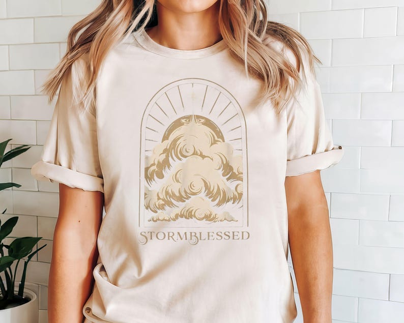 May include: A beige t-shirt with a graphic of a stylized cloud with a sun and rays behind it. The text "STORMBLESSED" is printed below the graphic.
