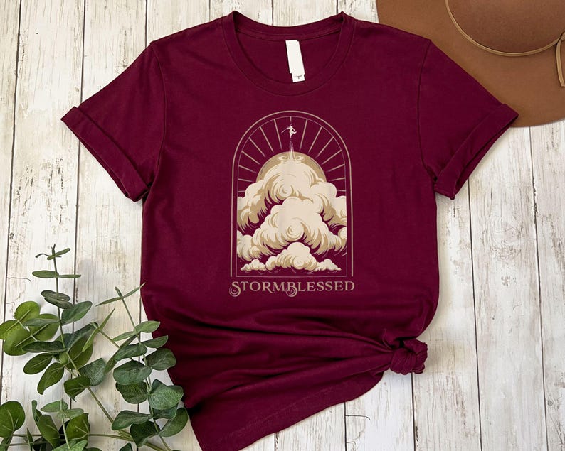 May include: A maroon t-shirt with a graphic of a sun, clouds, and a bird flying above the clouds. The text "STORMBLESSED" is printed below the graphic.