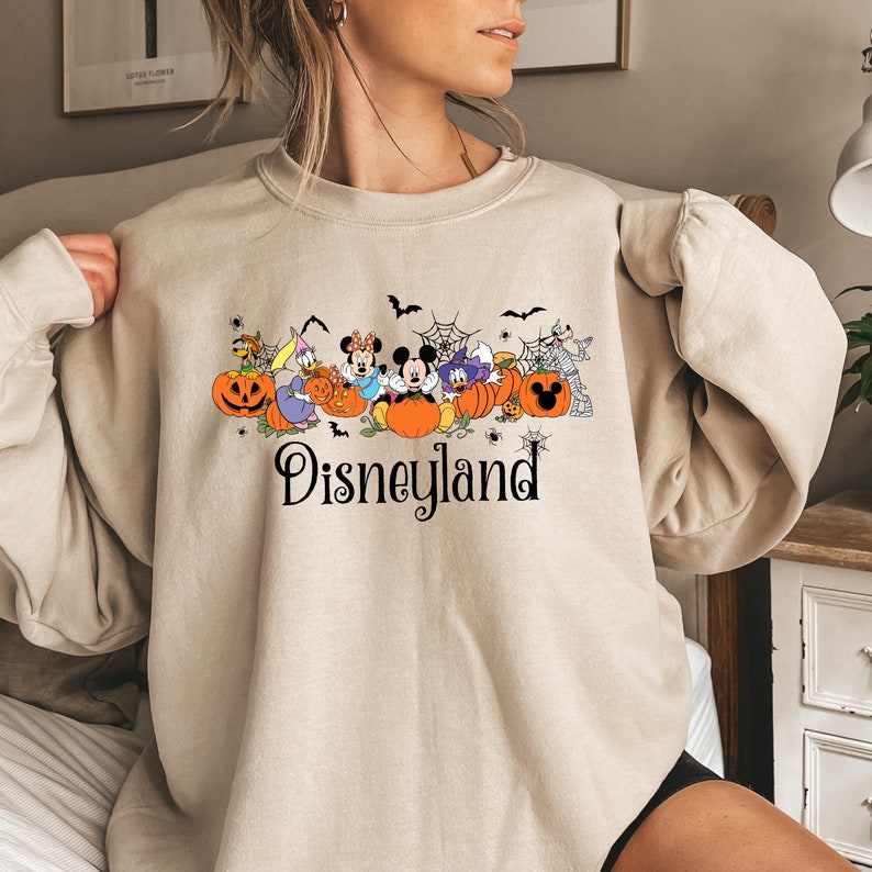 May include: A beige sweatshirt with a graphic print of Mickey Mouse and friends dressed in Halloween costumes. The graphic includes pumpkins, bats, and a spider web. The text "Disneyland" is printed below the graphic.