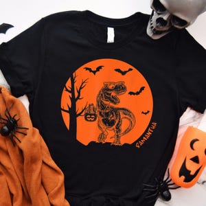 May include: Black t-shirt with an orange graphic of a dinosaur holding a jack-o-lantern in front of a full moon with bats flying around it. The text "SAMANTHA" is printed on the shirt.