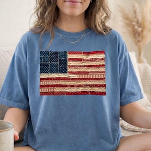 May include: A blue t-shirt featuring a distressed American flag design. The flag graphic is a patchwork of red, white, and blue fabrics, creating a vintage, handcrafted aesthetic. The shirt is a relaxed fit.