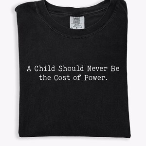 Puede incluir: Camiseta negra con el texto blanco "A Child Should Never Be the Cost of Power". La camiseta est&aacute; doblada cuidadosamente, mostrando el mensaje de forma prominente. La etiqueta Comfort Colors es visible en la parte superior.