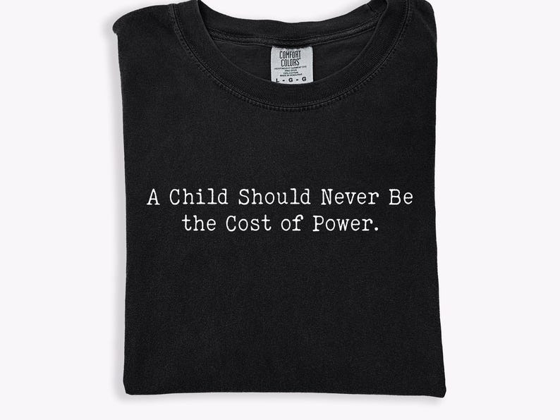 Puede incluir: Camiseta negra con el texto blanco "A Child Should Never Be the Cost of Power". La camiseta est&aacute; doblada cuidadosamente, mostrando el mensaje de forma prominente. La etiqueta Comfort Colors es visible en la parte superior.