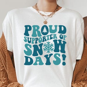 May include: White t-shirt with the text "PROUD SUPPORTER OF SNOW DAYS!" in teal and blue. The design includes a tree and snowflake graphic. The shirt is worn with a brown cardigan and gold necklace.