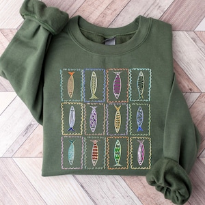 May include: A dark green sweatshirt with a colorful embroidered design of fish in squares. The fish are outlined in black and filled with various colors, including red, blue, green, yellow, and purple.
