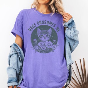 Puede incluir: Camiseta morada con un gr&aacute;fico de gato rodeado de flores dentro de un c&iacute;rculo. El texto "RAGE CONSUMES ME" est&aacute; arqueado encima del gato. La camiseta se usa con una chaqueta vaquera.