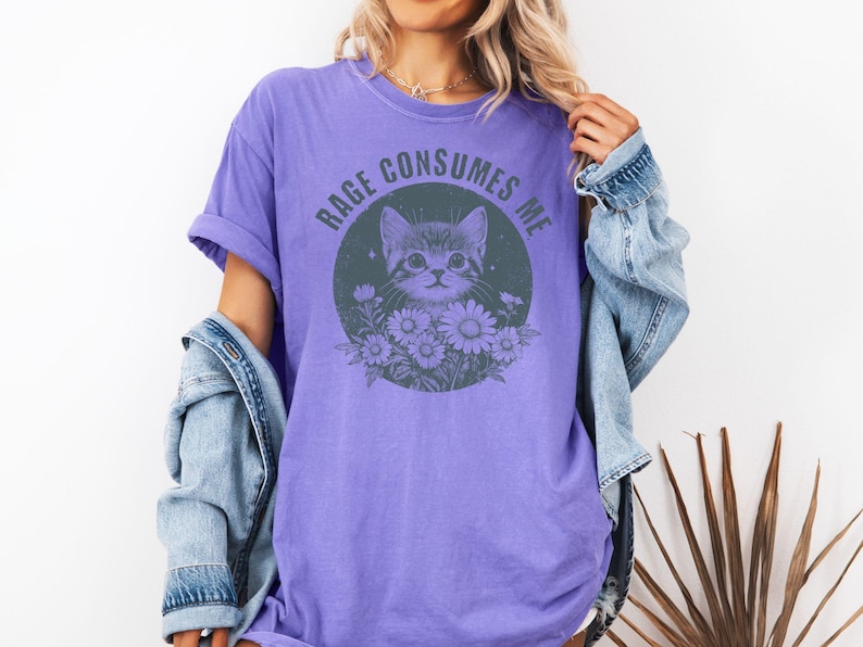 Puede incluir: Camiseta morada con un gr&aacute;fico de gato rodeado de flores dentro de un c&iacute;rculo. El texto "RAGE CONSUMES ME" est&aacute; arqueado encima del gato. La camiseta se usa con una chaqueta vaquera.