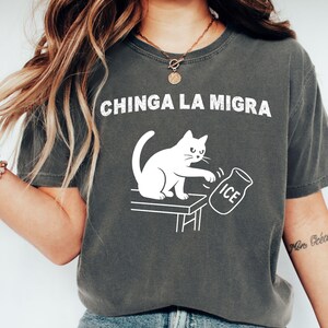 May include: Dark gray t-shirt with the text "CHINGA LA MIGRA" in white. A white cat is depicted pushing over a jar of ice. The shirt is a casual style, likely made of cotton or a similar fabric.