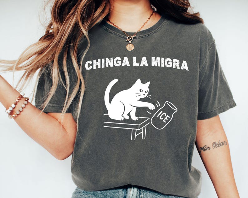 May include: Dark gray t-shirt with the text "CHINGA LA MIGRA" in white. A white cat is depicted pushing over a jar of ice. The shirt is a casual style, likely made of cotton or a similar fabric.