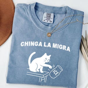 May include: A blue Comfort Colors t-shirt with the text "CHINGA LA MIGRA" and a white graphic of a cat knocking over a container of ice. A gold necklace and a brown hat are also visible.