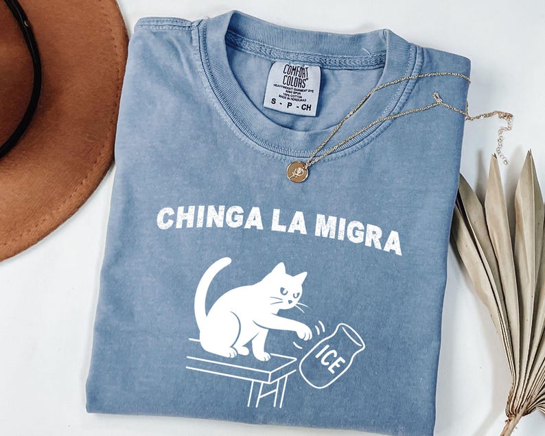 May include: A blue Comfort Colors t-shirt with the text "CHINGA LA MIGRA" and a white graphic of a cat knocking over a container of ice. A gold necklace and a brown hat are also visible.