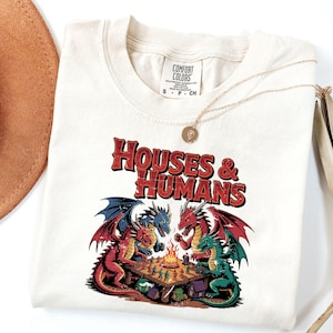 May include: Cream-colored t-shirt with the text "HOUSES & HUMANS" in red and black. The shirt features a graphic of four dragons playing a game around a campfire. A gold necklace is draped across the shirt.