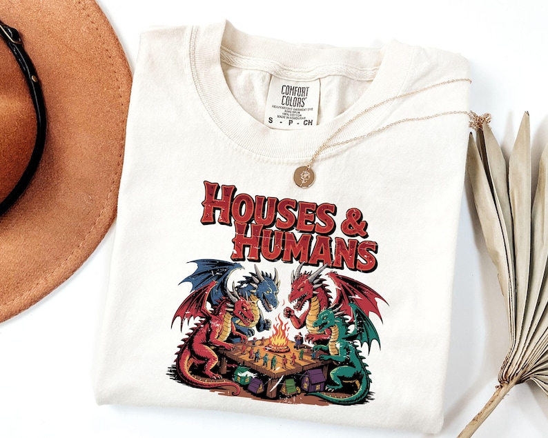 May include: Cream-colored t-shirt with the text "HOUSES & HUMANS" in red and black. The shirt features a graphic of four dragons playing a game around a campfire. A gold necklace is draped across the shirt.