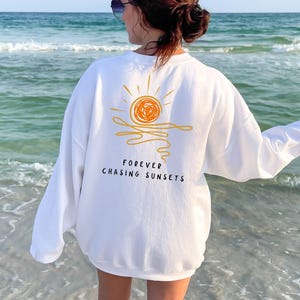 May include: White sweatshirt with a sunset graphic in orange and yellow. The text "FOREVER CHASING SUNSETS" is printed below the graphic. The sweatshirt is long-sleeved and the image is taken at the beach.