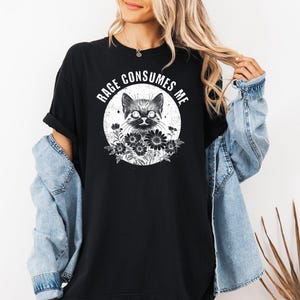Puede incluir: Camiseta negra con una ilustraci&oacute;n de un gato dentro de un c&iacute;rculo, rodeado de flores. El texto "RAGE CONSUMES ME" est&aacute; arqueado sobre el gato. La camiseta se lleva con una chaqueta vaquera.