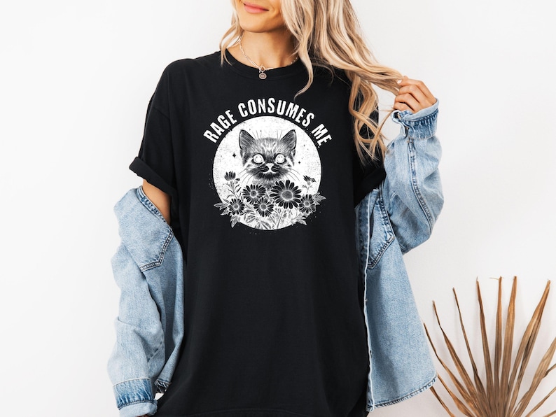 Puede incluir: Camiseta negra con una ilustraci&oacute;n de un gato dentro de un c&iacute;rculo, rodeado de flores. El texto "RAGE CONSUMES ME" est&aacute; arqueado sobre el gato. La camiseta se lleva con una chaqueta vaquera.