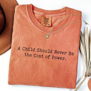 Puede incluir: Camiseta de color coral con el texto "A Child Should Never Be the Cost of Power". Un collar dorado con un colgante descansa sobre la camisa. Un sombrero marr&oacute;n y una planta seca est&aacute;n en el fondo.