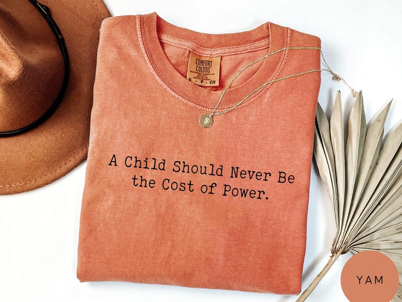 Puede incluir: Camiseta de color coral con el texto "A Child Should Never Be the Cost of Power". Un collar dorado con un colgante descansa sobre la camisa. Un sombrero marr&oacute;n y una planta seca est&aacute;n en el fondo.