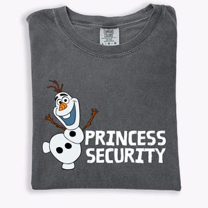 May include: Dark gray t-shirt featuring a cartoon snowman character with the text "PRINCESS SECURITY" in white. The snowman has a carrot nose, twig arms, and a smiling expression. The shirt is folded and has a Comfort Colors tag.