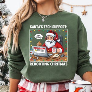 May include: Green sweatshirt with a graphic of Santa Claus wearing headphones and working on a laptop. The text reads "SANTA'S TECH SUPPORT: REBOOTING CHRISTMAS." Santa is holding a cookie and a mug that says "#1 Support Elf."