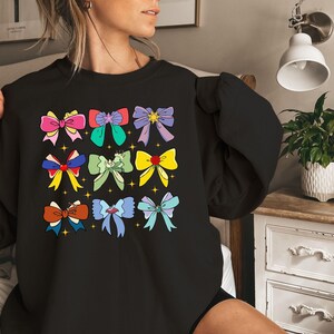 May include: Black sweatshirt with a colorful bow pattern. The bows are in various colors, including pink, blue, green, yellow, and purple. Each bow has a gold star in the center.