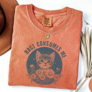 Puede incluir: Camiseta de color naranja rojizo con el texto "RAGE CONSUMES ME" que rodea una ilustraci&oacute;n de un gato. El gato est&aacute; en un c&iacute;rculo azul oscuro con flores. Un collar dorado est&aacute; sobre la camiseta. Un sombrero marr&oacute;n es visible en la esquina superior izquierda.