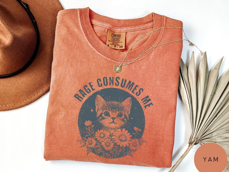 Puede incluir: Camiseta de color naranja rojizo con el texto "RAGE CONSUMES ME" que rodea una ilustraci&oacute;n de un gato. El gato est&aacute; en un c&iacute;rculo azul oscuro con flores. Un collar dorado est&aacute; sobre la camiseta. Un sombrero marr&oacute;n es visible en la esquina superior izquierda.