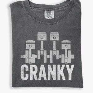 May include: Dark gray t-shirt with a white graphic of four pistons and the word "CRANKY" in a distressed font. The shirt is folded and has a Comfort Colors tag.