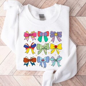 May include: White sweatshirt with a colorful bow design. The bows are in various colors, including pink, blue, green, yellow, and red. Each bow has a different design, including stars, hearts, and swirls.