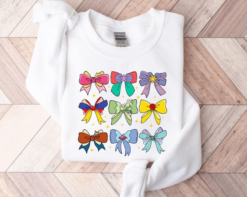 May include: White sweatshirt with a colorful bow design. The bows are in various colors, including pink, blue, green, yellow, and red. Each bow has a different design, including stars, hearts, and swirls.