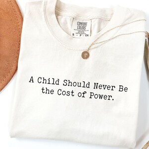Puede incluir: Camiseta color crema con el texto "A Child Should Never Be the Cost of Power". Un collar dorado con un peque&ntilde;o colgante descansa sobre la camiseta. Un sombrero marr&oacute;n y una planta seca est&aacute;n en el fondo.