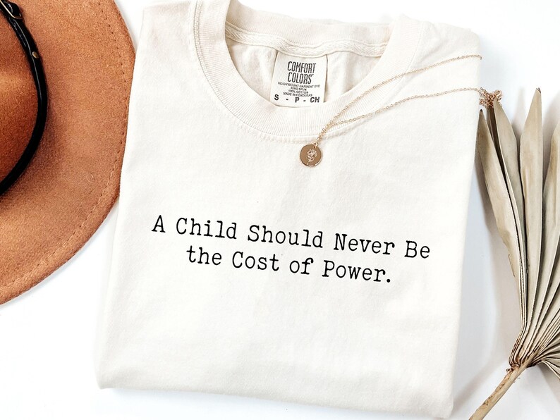 Puede incluir: Camiseta color crema con el texto "A Child Should Never Be the Cost of Power". Un collar dorado con un peque&ntilde;o colgante descansa sobre la camiseta. Un sombrero marr&oacute;n y una planta seca est&aacute;n en el fondo.