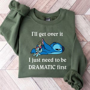 May include: Green sweatshirt featuring a blue Stitch character from Lilo & Stitch. The text on the sweatshirt reads "I'll get over it I just need to be DRAMATIC first."