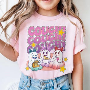 Puede incluir: Camiseta rosa con la frase "COUCH! COUCH! COUCH!" en una fuente retro. Debajo, tres fantasmas de dibujos animados con lazos, sosteniendo bocadillos. El fantasma de la izquierda sostiene una bolsa de patatas fritas "POTATO".