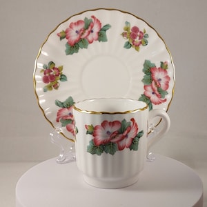 Può includere: Una tazza da tè e un piattino bianchi con un motivo floreale. La tazza e il piattino hanno un bordo dorato e presentano fiori rosa e verdi.