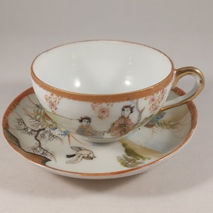 May include: A white porcelain teacup and saucer set with a gold rim and a hand-painted design of Japanese figures in a garden setting.
