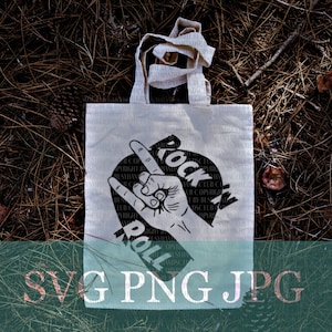 May include: A cream-colored tote bag with a black graphic of a hand making the rock and roll sign. The words "Rock" and "Roll" are printed on the bag. The bag has a handle and is sitting on a bed of pine needles.